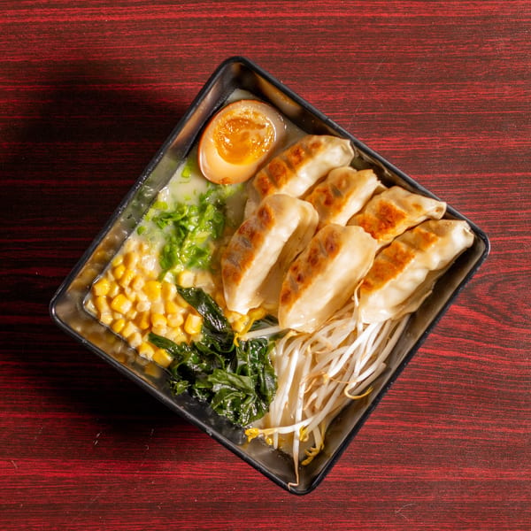 A square bowl of ramen with gyoza, corn, seaweed, bean sprouts, and a soft-boiled egg.