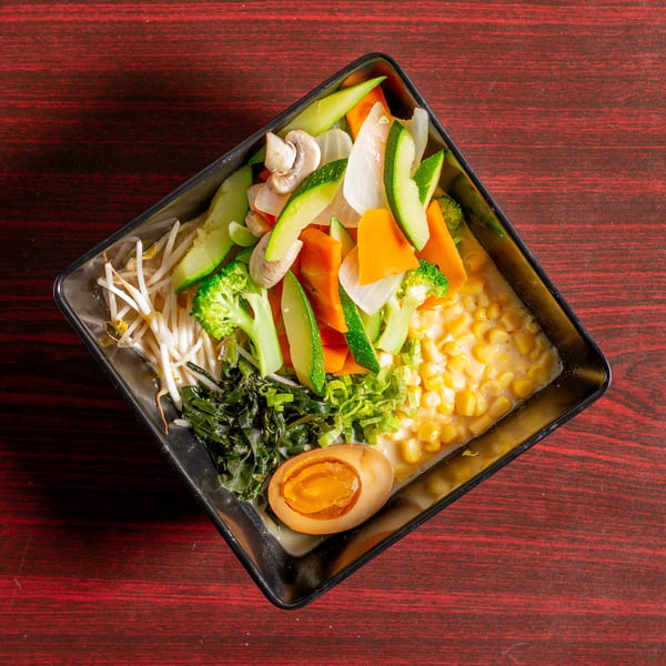A square bowl of ramen with various vegetables, corn, seaweed, and a soft-boiled egg.